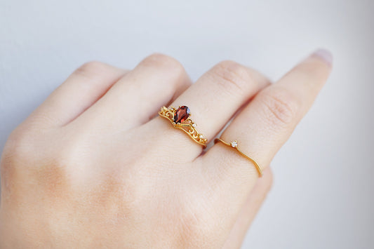 Gold ring with a red gemstone on a finger against a light gray background