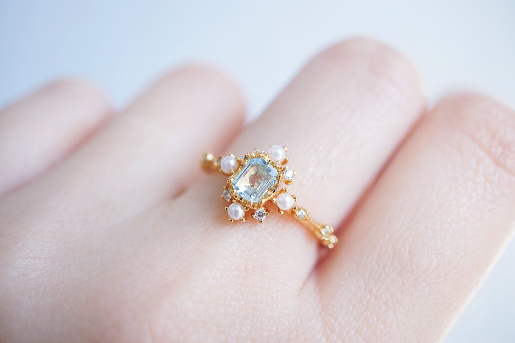 Gold ring with a central gemstone and pearls on a finger against a light background