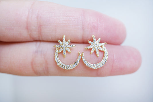 Gold earrings with star and crescent moon designs held between fingers on a light background