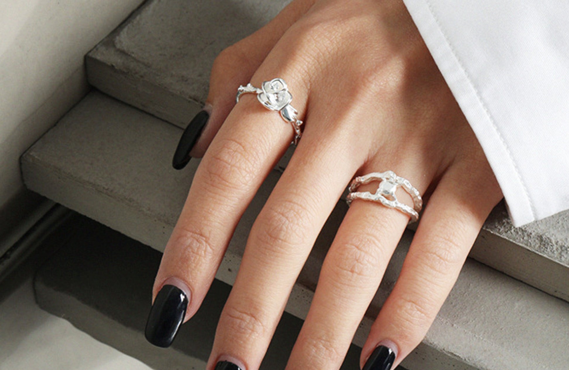 Hand wearing two silver rings with black nail polish on a neutral background