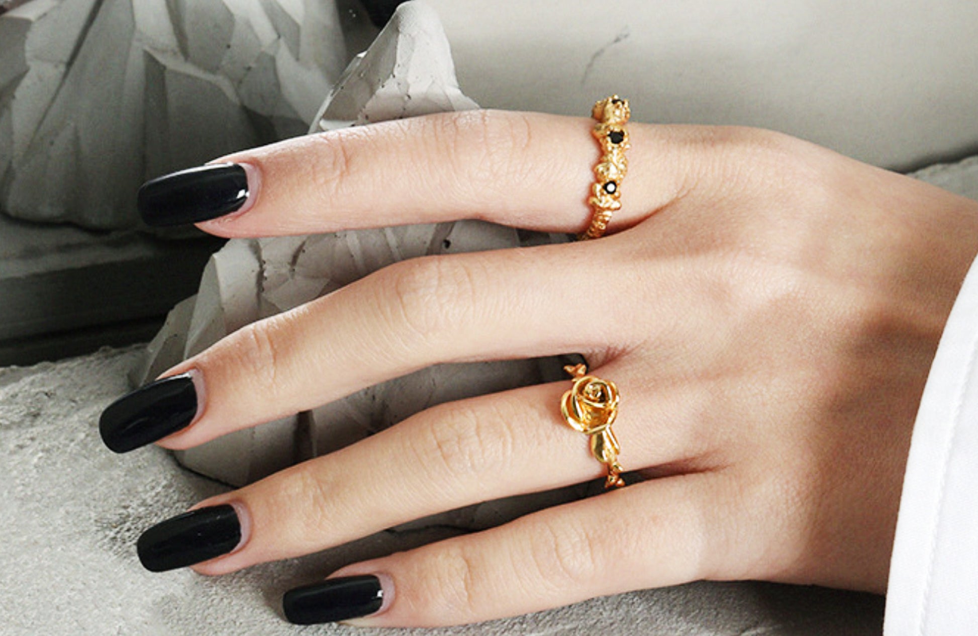 Hand wearing two gold rings with black nail polish on a neutral background