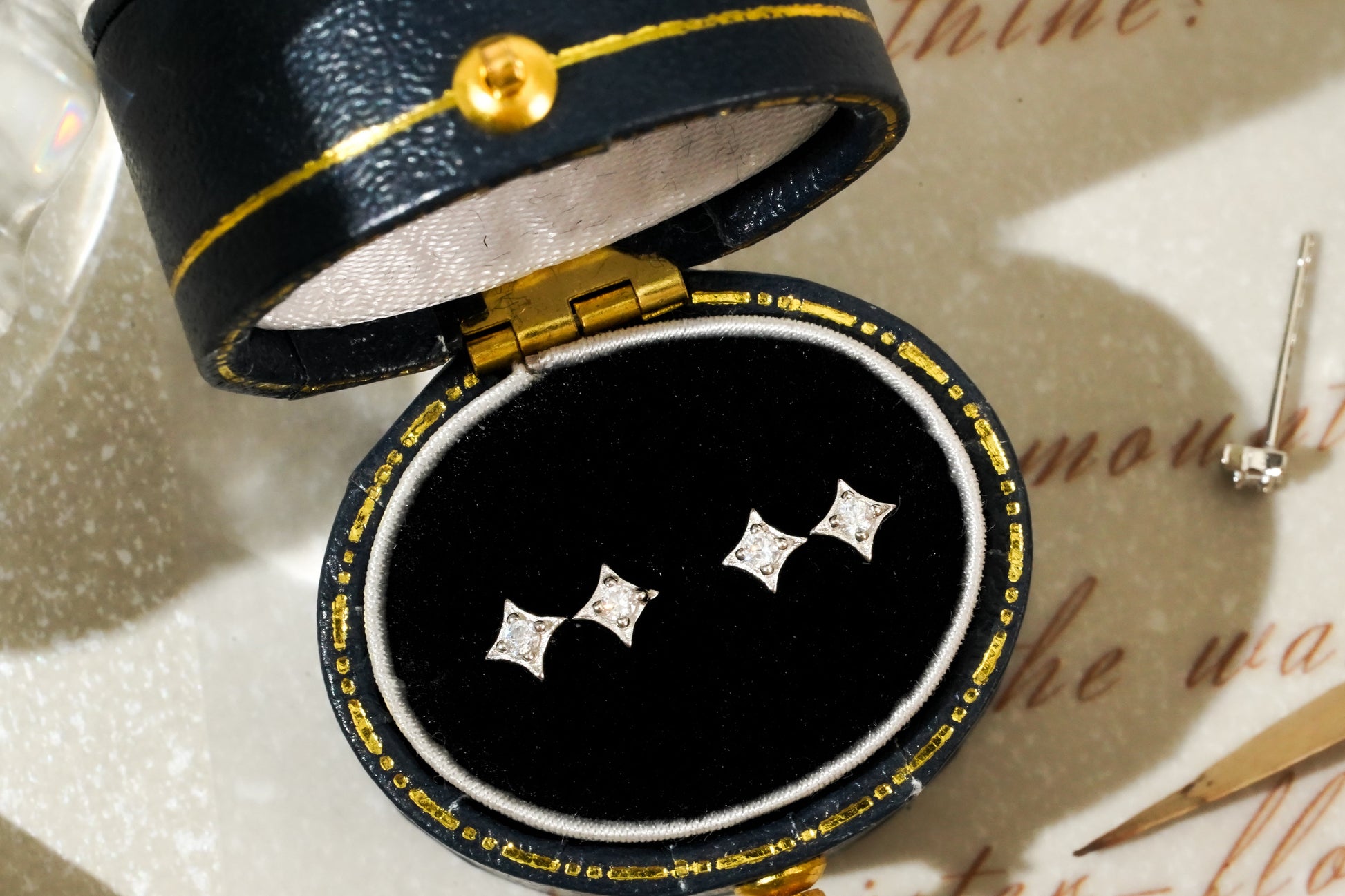 Black jewelry box with gold accents containing diamond earrings on a textured surface.